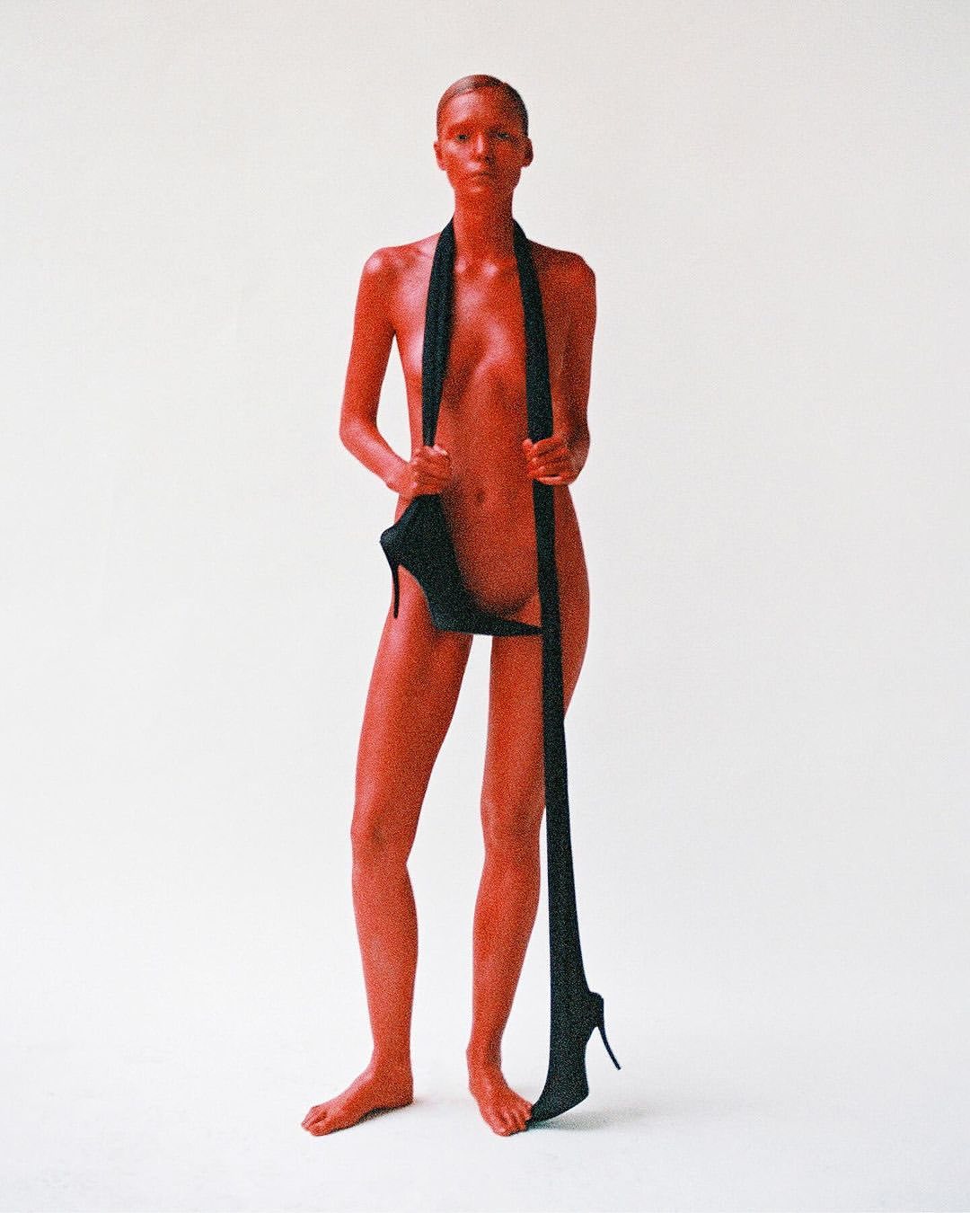 Adult female standing in a bright white photo studio, with high heel shoes suspended from her neck.