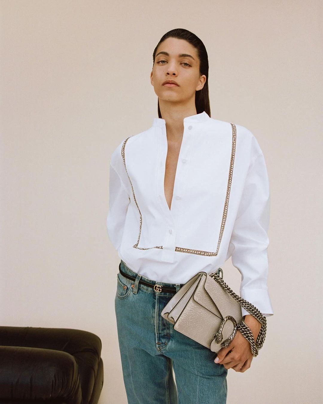 An adult female poses in a white button-down shirt, light-wash jeans, and a neutral-colored handbag with a chain strap in a studio setting.