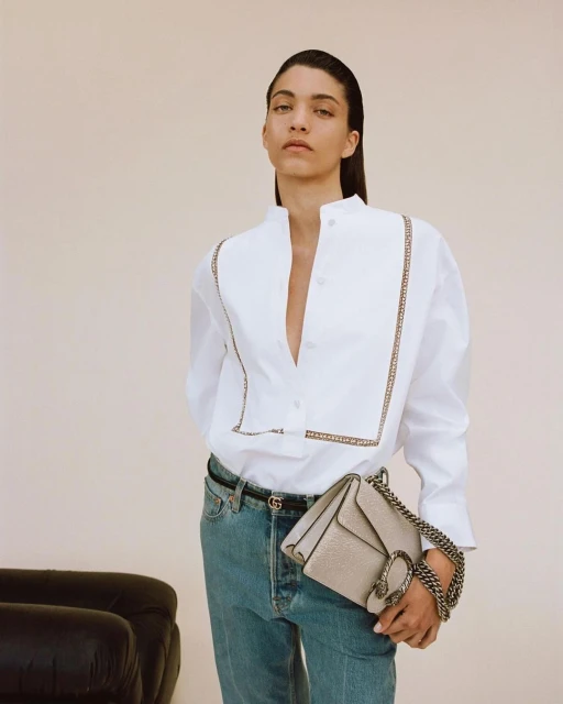 An adult female poses in a white button-down shirt, light-wash jeans, and a neutral-colored handbag with a chain strap in a studio setting.