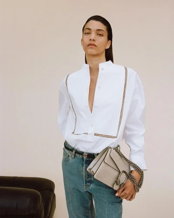 An adult female poses in a white button-down shirt, light-wash jeans, and a neutral-colored handbag with a chain strap in a studio setting.