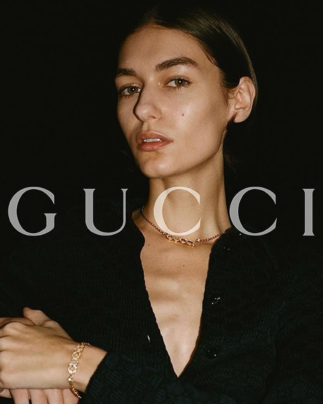 Close-up fashion shot of a female model wearing a black cardigan and gold jewelry against a dark background with brand logo.