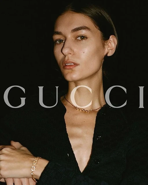 Close-up fashion shot of a female model wearing a black cardigan and gold jewelry against a dark background with brand logo.