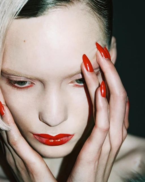 Close-up shot of a fair-skinned woman with striking red lipstick and long, red stiletto nails, holding her hands near her face in a beauty-focused image.