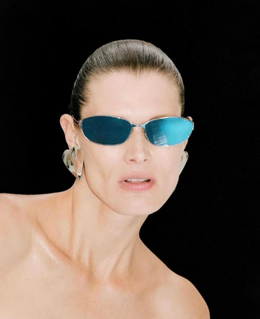Close-up of an adult woman with slicked-back hair wearing silver sunglasses and heart-shaped earrings against a dark background.