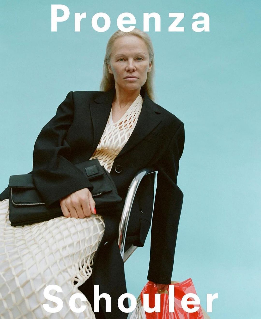 Woman sits in a black blazer and cream dress while holding a black purse and plastic shopping bag in a fashion editorial studio shot.