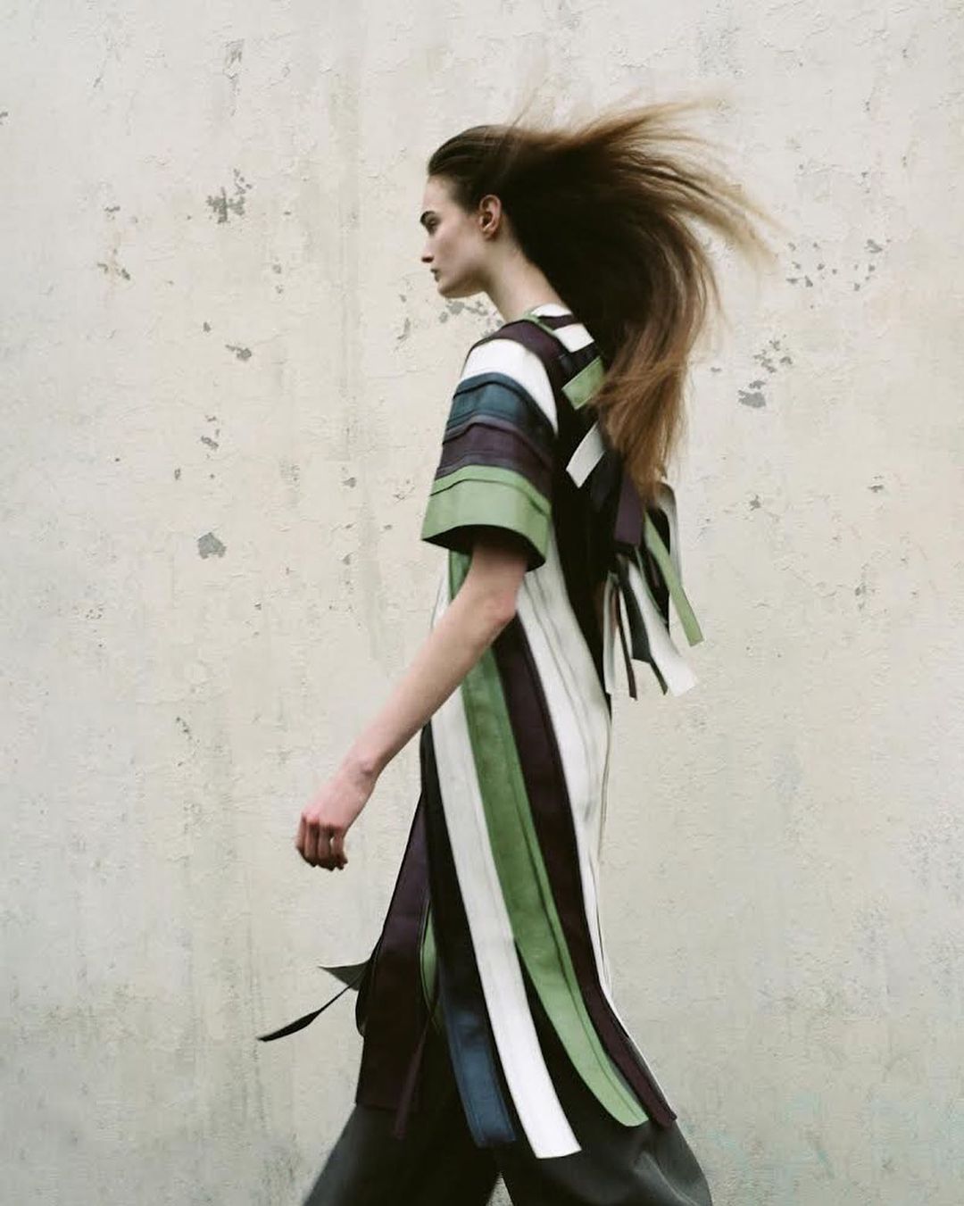 Model in profile with windblown hair walking past a textured distressed concrete wall wearing a striped tunic dress.