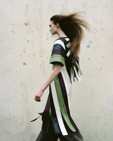 Model in profile with windblown hair walking past a textured distressed concrete wall wearing a striped tunic dress.