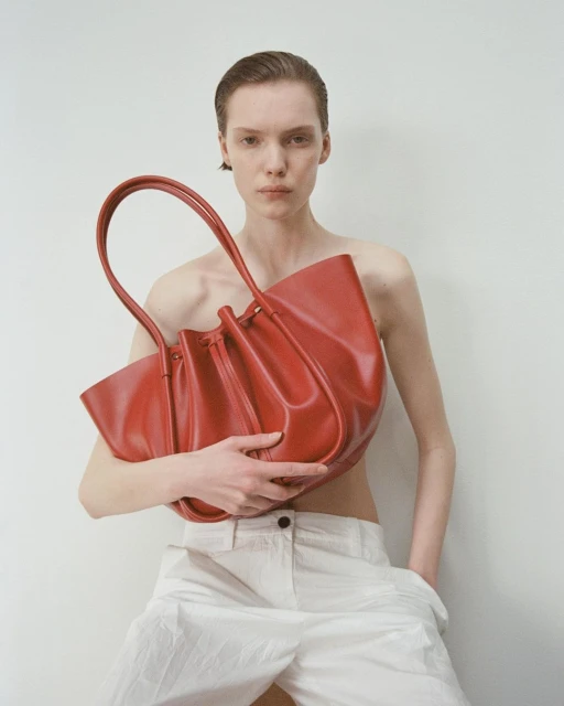 Fashion shot of a model holding a large red leather tote bag, wearing white pants against a plain white studio background with soft lighting.