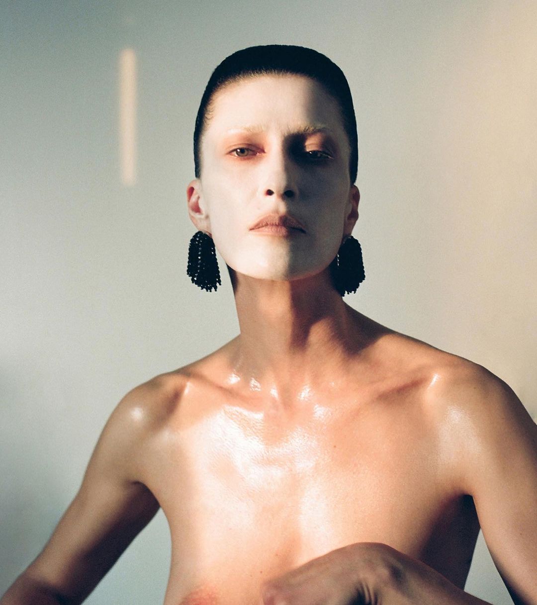 Close-up studio shot of androgynous model with pale skin, slicked-back hair, and large black tassel earrings; a stark and striking beauty image.