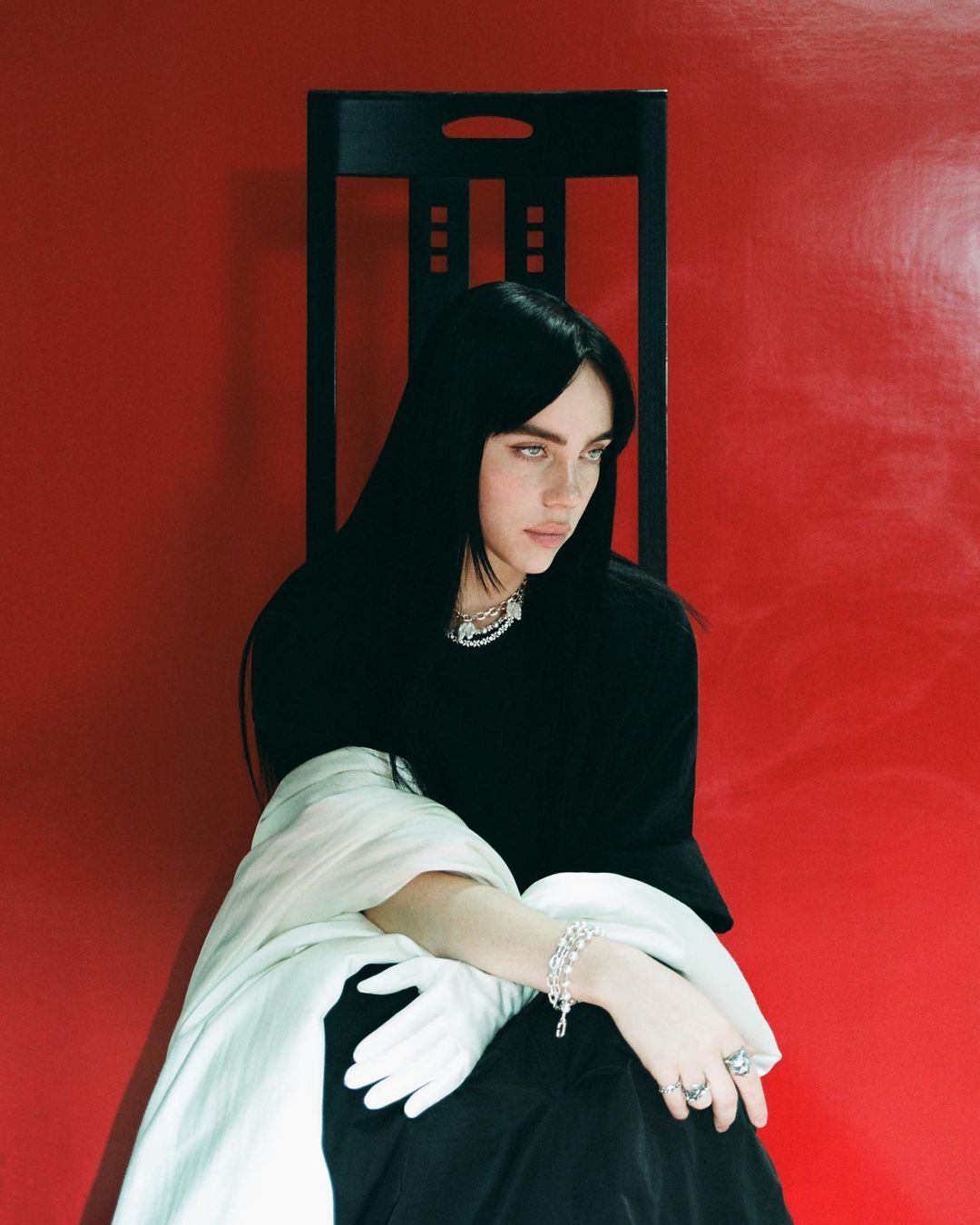 Portrait of a young adult woman with long, dark hair, seated in front of a red wall with a black chair, wearing jewelry and gloves.