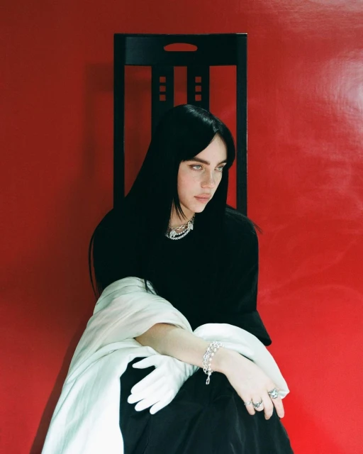 Portrait of a young adult woman with long, dark hair, seated in front of a red wall with a black chair, wearing jewelry and gloves.
