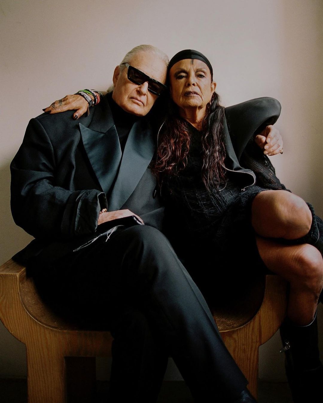 Two older adults in dark attire pose indoors. The man wears sunglasses and a suit; the woman, a dark dress and head covering.