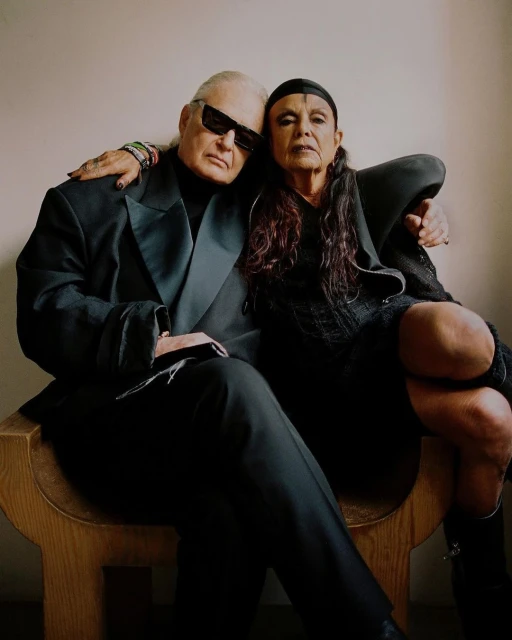 Two older adults in dark attire pose indoors. The man wears sunglasses and a suit; the woman, a dark dress and head covering.