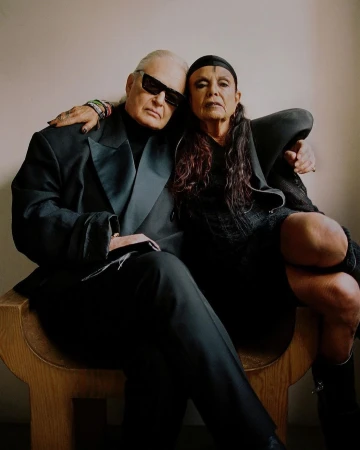 Two older adults in dark attire pose indoors. The man wears sunglasses and a suit; the woman, a dark dress and head covering.
