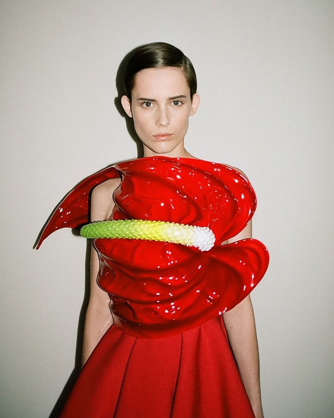 Elegant adult female poses indoors wearing a unique red sculptural top and matching A-line skirt, against a neutral wall backdrop.