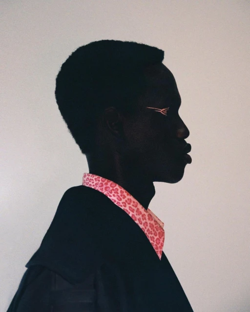 Profile of a dark-skinned person with pink eyeliner and a pink cheetah-print collared shirt, wearing a black jacket against an off-white background.