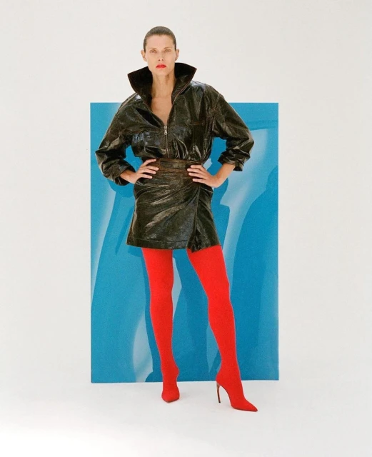 A fair-skinned woman poses in a dark jacket and skirt with bright red tights, standing against a blue and white backdrop in a studio.