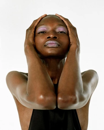 Close-up studio portrait of an African female with hands framing her face, featuring dramatic lavender makeup on her eyes and lips.