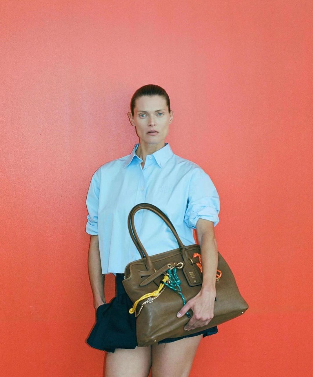 Woman posing with a tan leather satchel handbag, wearing a light blue cropped top and dark shorts against a coral colored wall.