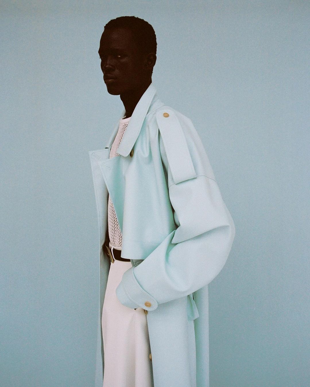 Fashion shot of a man wearing a light blue coat over a knitted white top and white pants, posing against a light blue background.