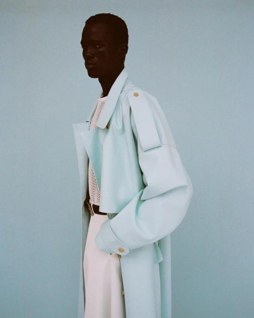 Fashion shot of a man wearing a light blue coat over a knitted white top and white pants, posing against a light blue background.