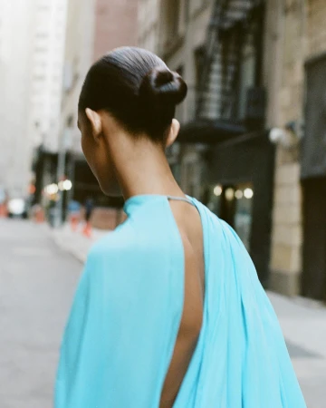 Model with slicked-back hair in a bright blue dress walking down a city street in New York, captured from behind.