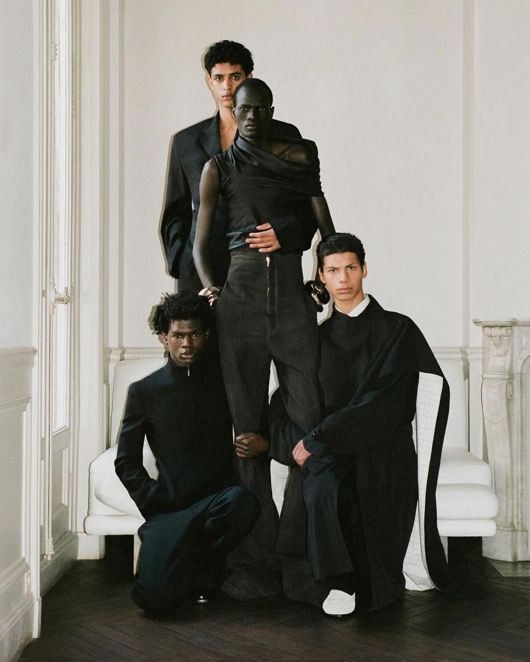 Four adult male fashion models are posing indoors against a white wall and white couch, all are wearing black clothing in studio light.