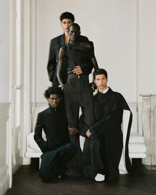 Four adult male fashion models are posing indoors against a white wall and white couch, all are wearing black clothing in studio light.