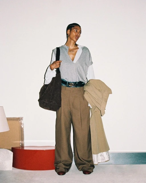 A stylish, full-length studio shot features a Black woman in an oversized grey top, brown trousers, and red shoes holding a jacket.
