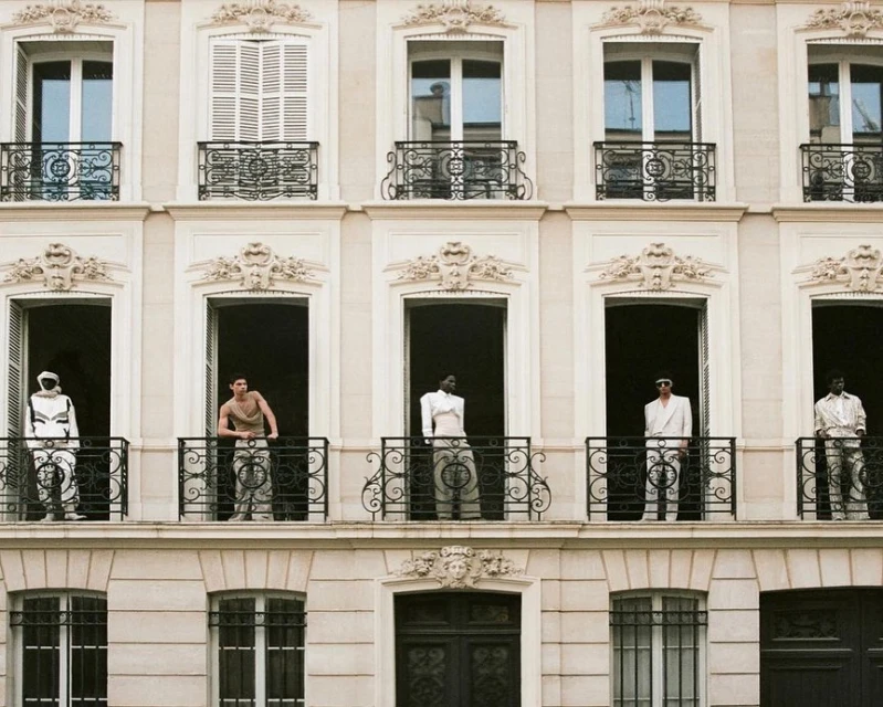 Five fashion models in architectural windows with wrought-iron balconies wear designer clothing. Light beige building facade in daylight.