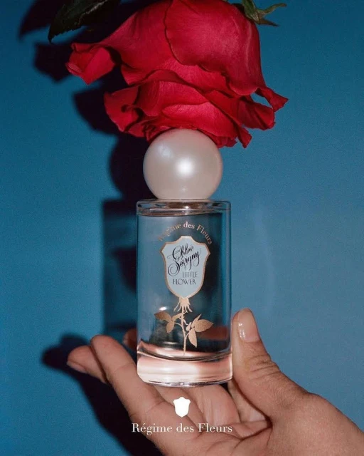 Perfume bottle topped with a rose, held in hand against a blue backdrop, product shot for Régime des Fleurs, beauty, fashion, still life.
