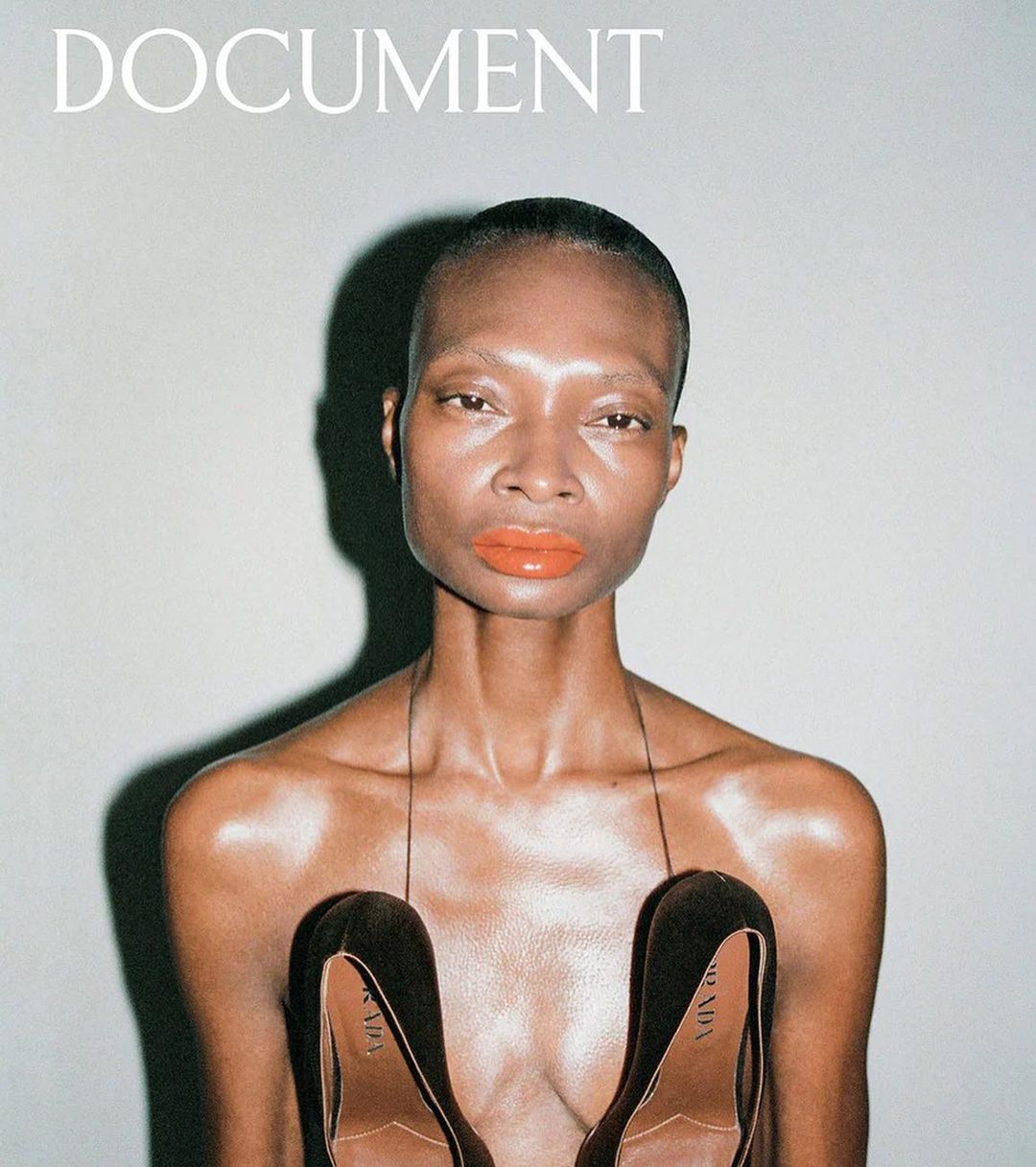 Fashion magazine cover featuring a woman with orange lipstick and black heels held at the chest, on a grey background.