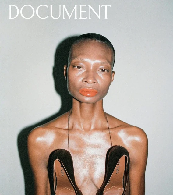 Fashion magazine cover featuring a woman with orange lipstick and black heels held at the chest, on a grey background.