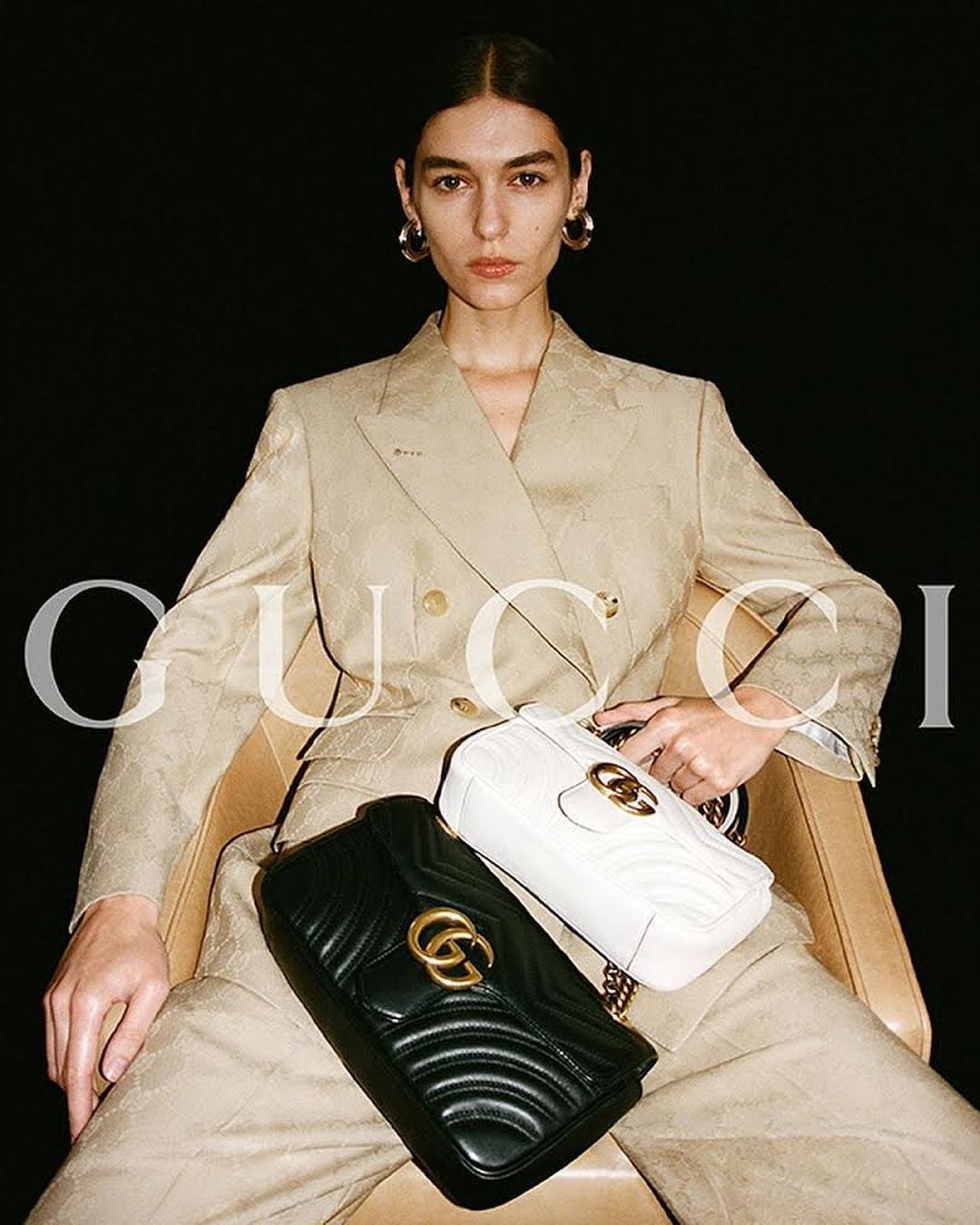 Model seated wearing a beige blazer and trousers holding two quilted leather handbags against a dark background
