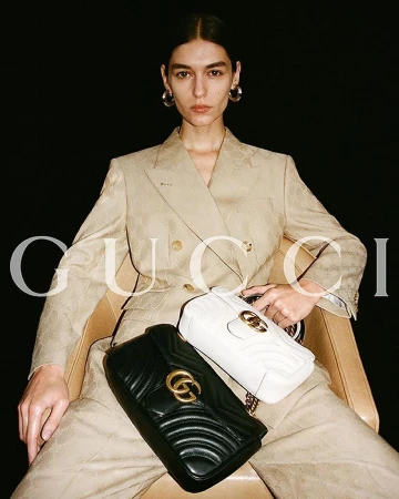Model seated wearing a beige blazer and trousers holding two quilted leather handbags against a dark background