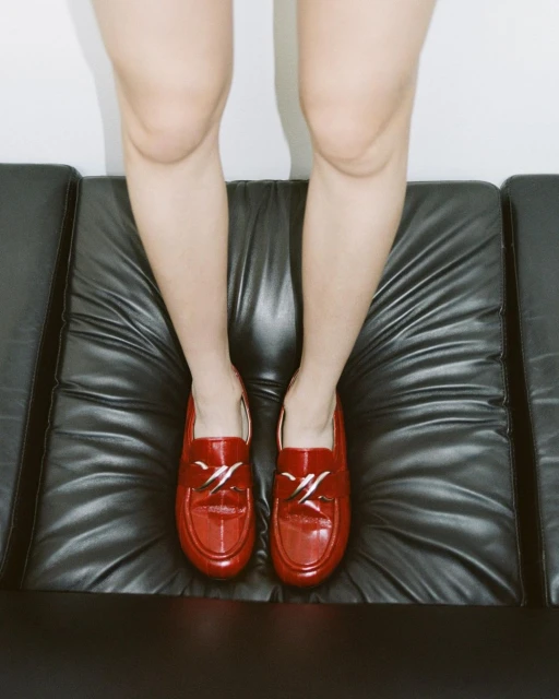 Close-up shot of person's legs and feet wearing red patent leather loafers while sitting on black leather cushioned bench against white wall.