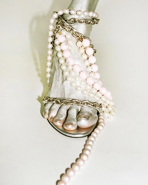 Close-up of a metallic-painted foot wearing a silver sandal, adorned with pearls and a silver chain, against a white background.