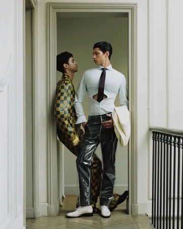 Two adult men in a doorway, one wearing a yellow-black checkerboard suit, the other wearing plastic pants, a partially unbuttoned shirt, and tie.
