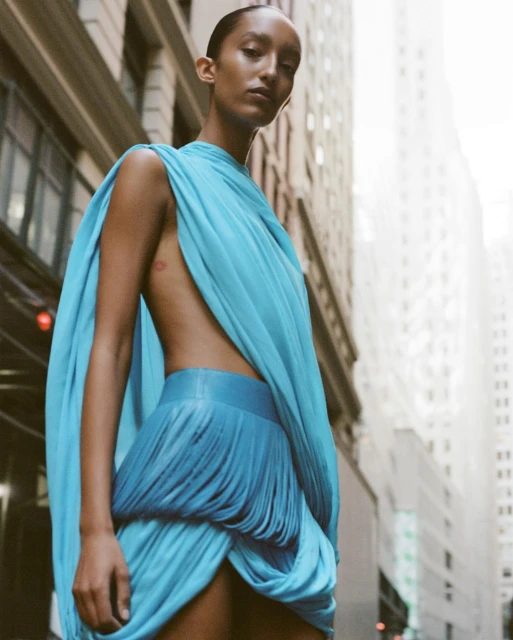 Elegant adult female model in a draped blue dress poses in an urban setting with tall buildings visible in the background.
