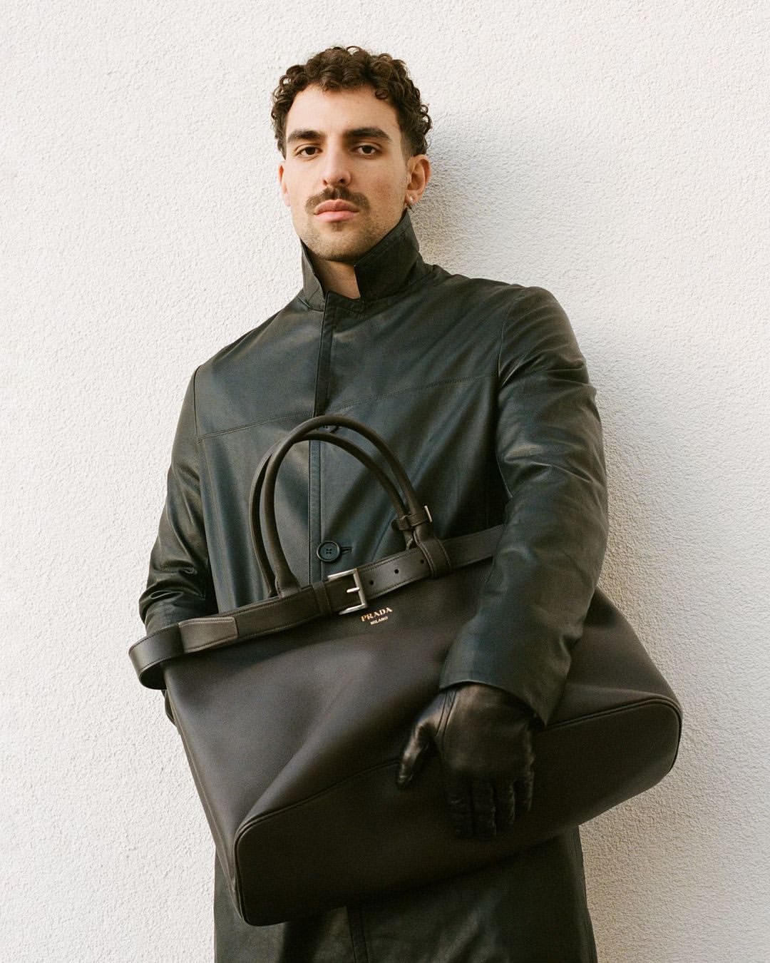 Man in a black coat and leather gloves holding a black Prada tote bag against a white wall in a portrait-style fashion shot.
