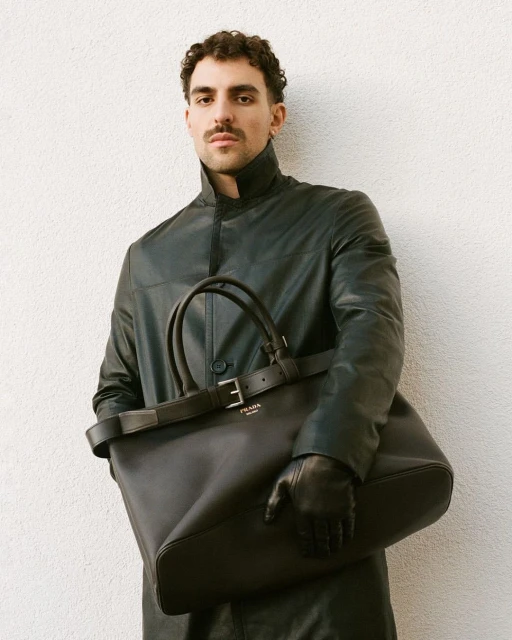 Man in a black coat and leather gloves holding a black Prada tote bag against a white wall in a portrait-style fashion shot.