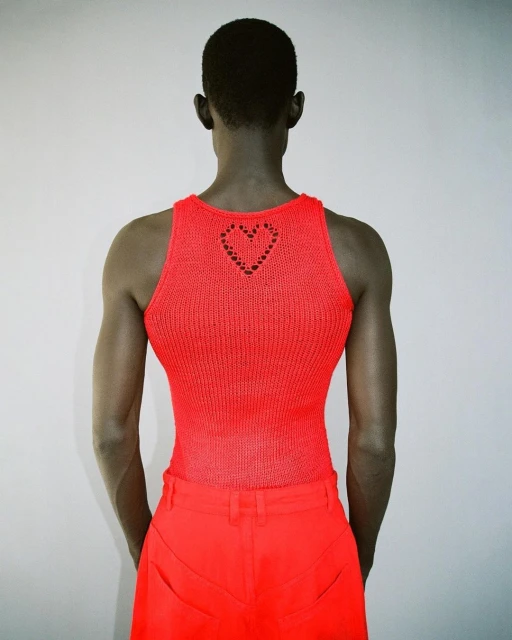 Back view of androgynous fashion model in coral tank top and pants with a heart cutout, posing against a light gray studio backdrop.