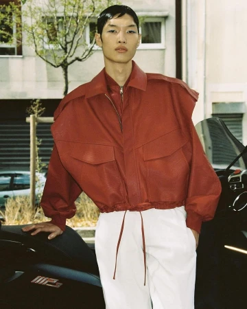 East Asian male model wearing a red jacket and white trousers standing outdoors by a vehicle