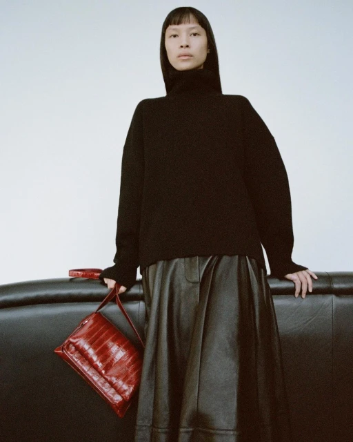 Asian woman in a black turtleneck and leather skirt holding a red crocodile-texture purse indoors against a white background.