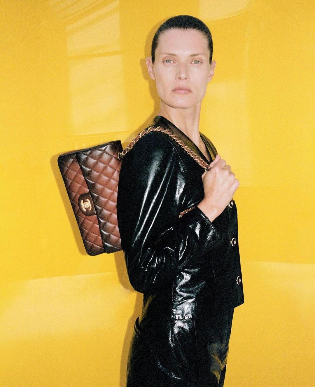 Fashion shot featuring an adult female model with slicked back hair, wearing a black leather jacket and skirt and a brown leather bag.