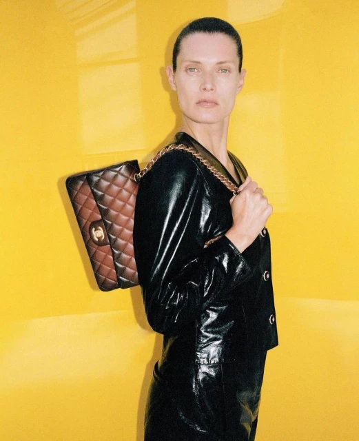 Fashion shot featuring an adult female model with slicked back hair, wearing a black leather jacket and skirt and a brown leather bag.