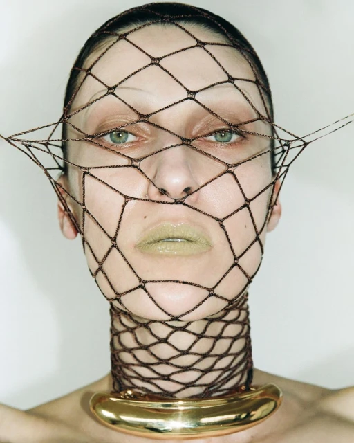 Close-up studio shot of a woman wearing a brown net face covering and gold choker. The focus is on the woman's face and the unique net.