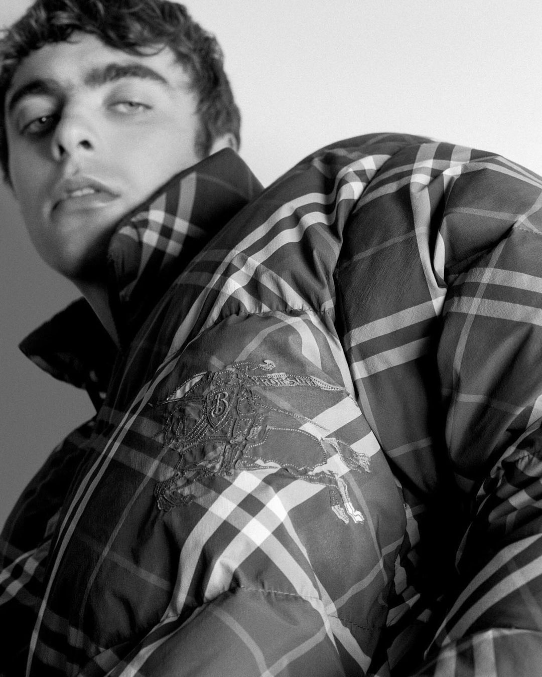 Monochrome portrait of a young adult male wearing a plaid puffer jacket, close-up shot against a white background, fashion-focused.