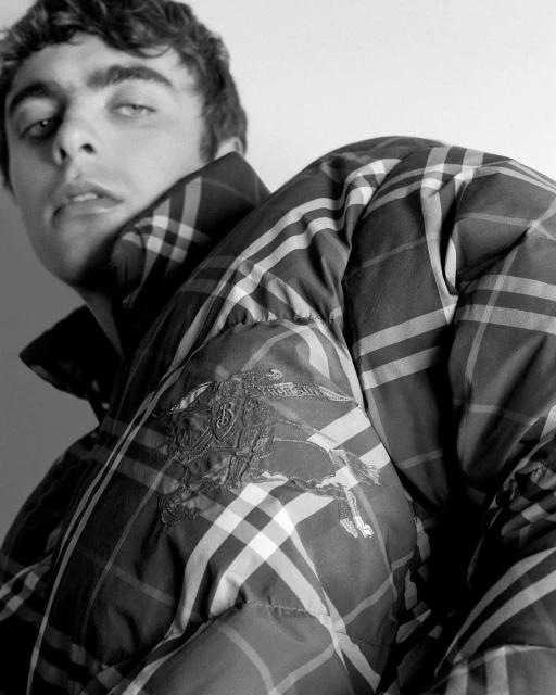 Monochrome portrait of a young adult male wearing a plaid puffer jacket, close-up shot against a white background, fashion-focused.