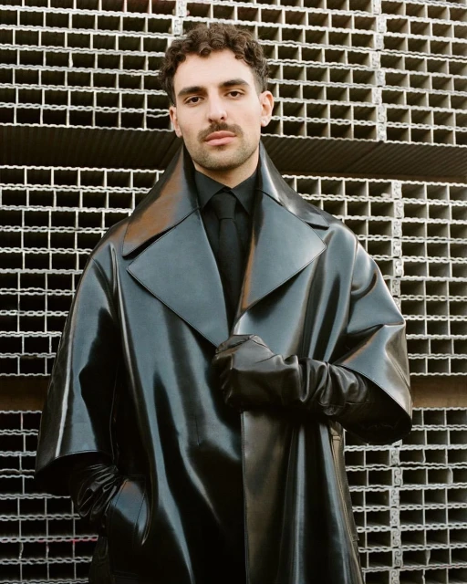 Portrait of a man in a black overcoat and black gloves standing against a metal grid backdrop, shot with natural lighting.